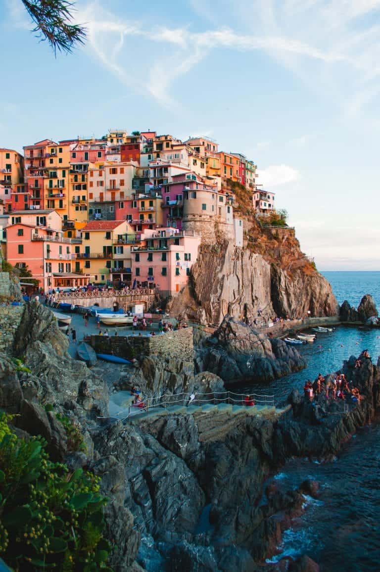 Experience the vibrant charm of this Italian coastal village, an ultimate bucket list destination. Perched on rocky cliffs, it boasts colorful facades and lively boats with people enjoying the clear sky's embrace by the water.