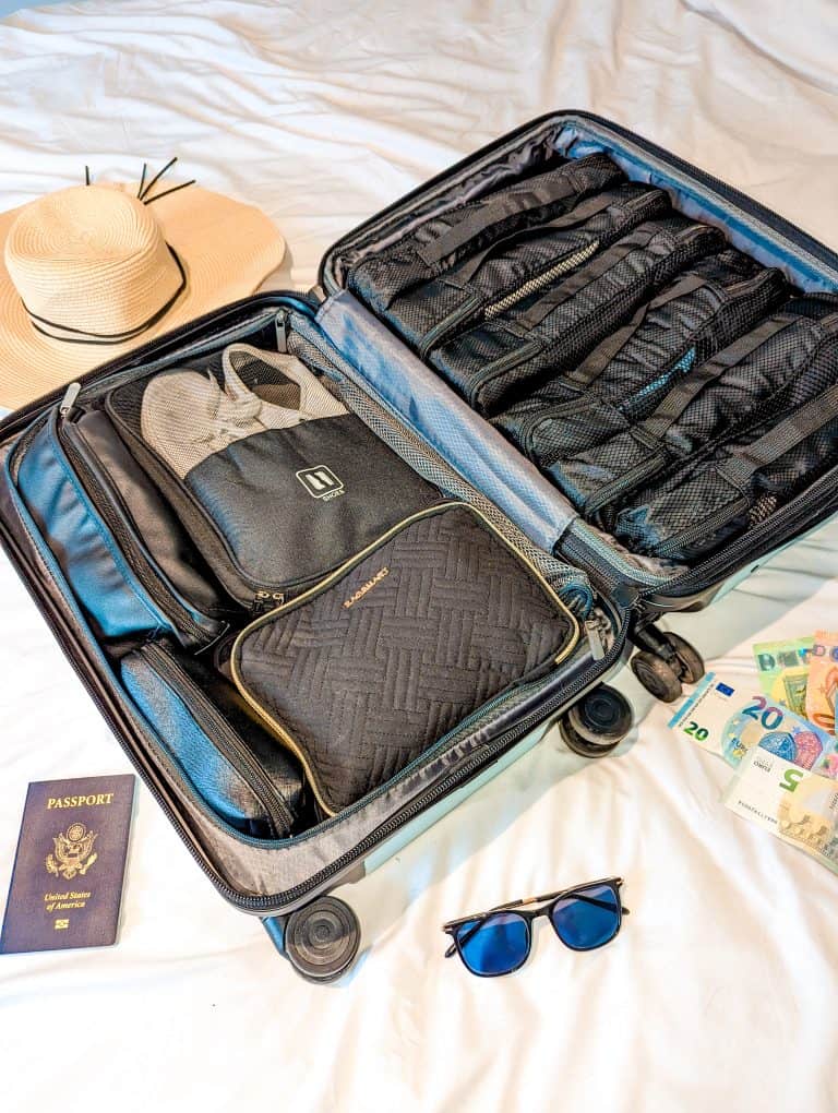 A suitcase with a hat, sunglasses, and money on a bed.
