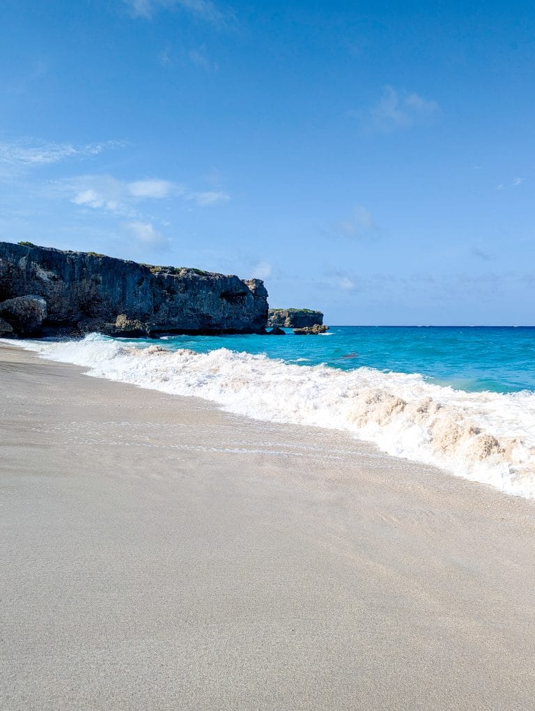 A sandy beach with waves and cliffs in the background.