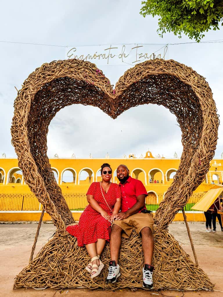 A couple sits on a heart shaped structure in front of a yellow building, fulfilling their Couples Travel Bucket List.