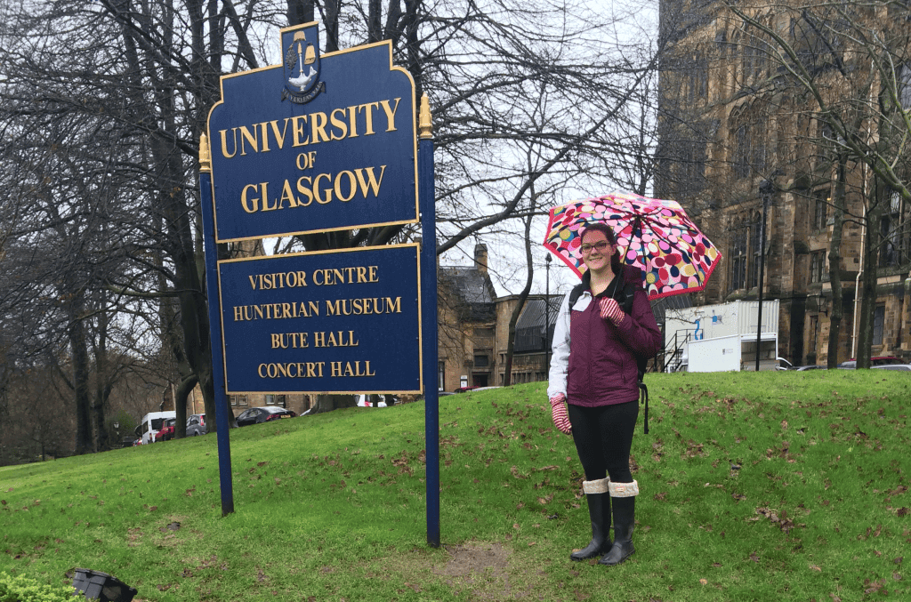 A woman holding an umbrella in front of a university of glasgow sign.