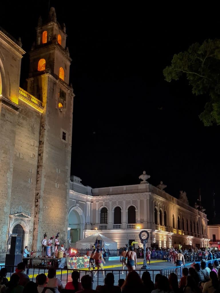 A vibrant nighttime scene in Merida with people near a historic building, illuminated by colorful lights.