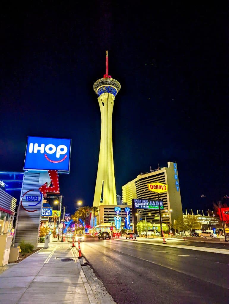 Night view of a brightly lit street with the stratosphere tower in the background and restaurant signs including ihop and denny's at the forefront.