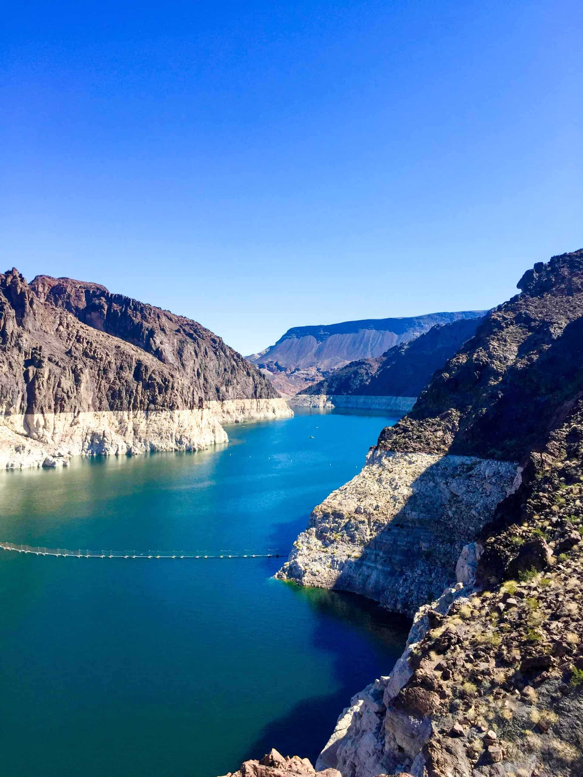 A serene view of a calm blue reservoir bordered by rugged mountains under a clear sky.