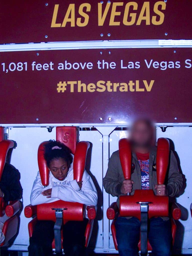 Two people strapped into a ride with a sign reading "las vegas 1,081 feet above the las vegas strip #thestratlv" in the background.