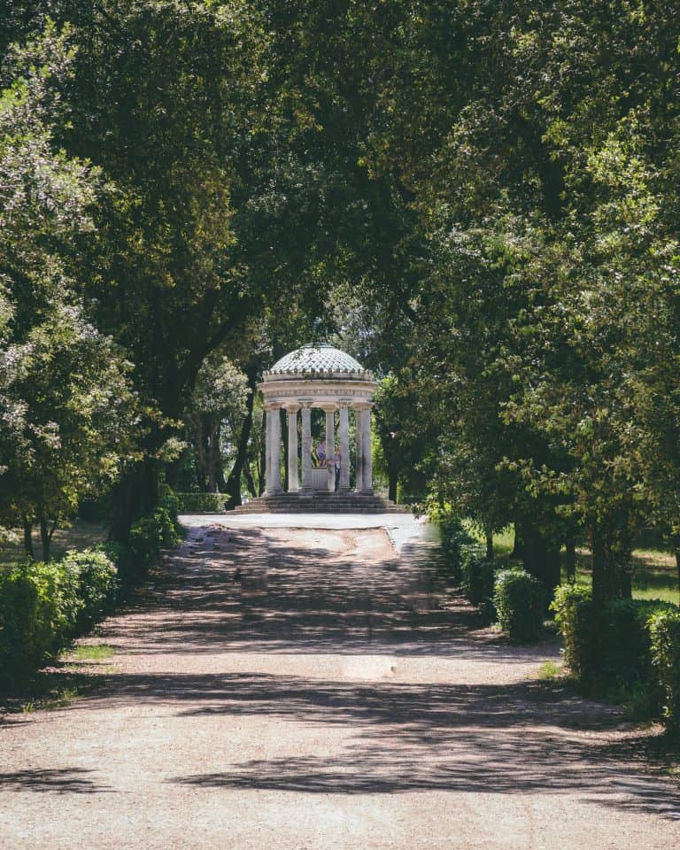 A stone gazebo with a dome roof, reminiscent of Rome's architectural charm, is surrounded by lush trees at the end of a sunlit path. It's a picturesque spot that deserves a place on your travel bucket list.