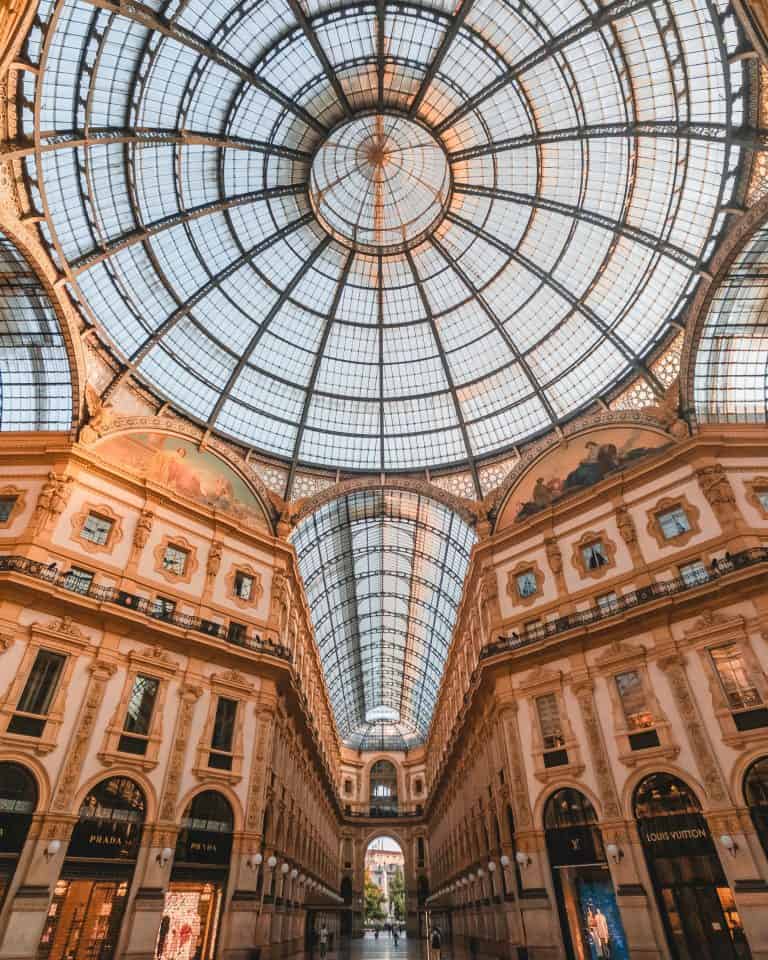 The interior of this grand shopping arcade, a Milan Bucket List essential, boasts a stunning glass dome ceiling and intricate architectural details. Illuminated storefronts elegantly line the walkway, offering an unparalleled shopping experience.