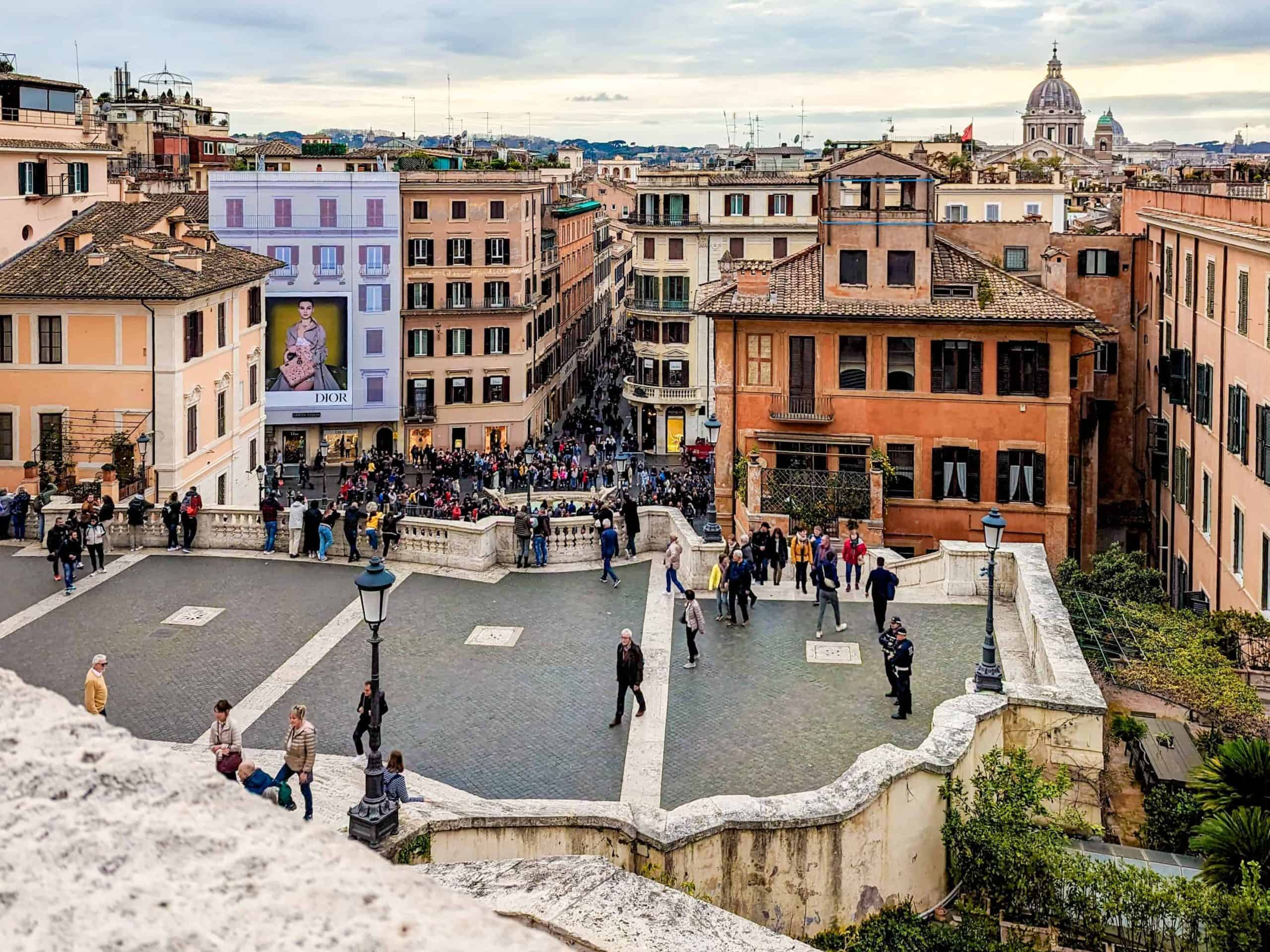 View of a bustling city square with historical buildings and a crowd of people, under an overcast sky.