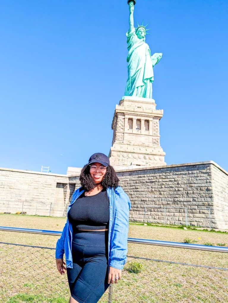 A woman in a black outfit and denim shirt stands smiling in front of the Statue of Liberty on a clear day, ticking off one iconic spot from her New York City bucket list.
