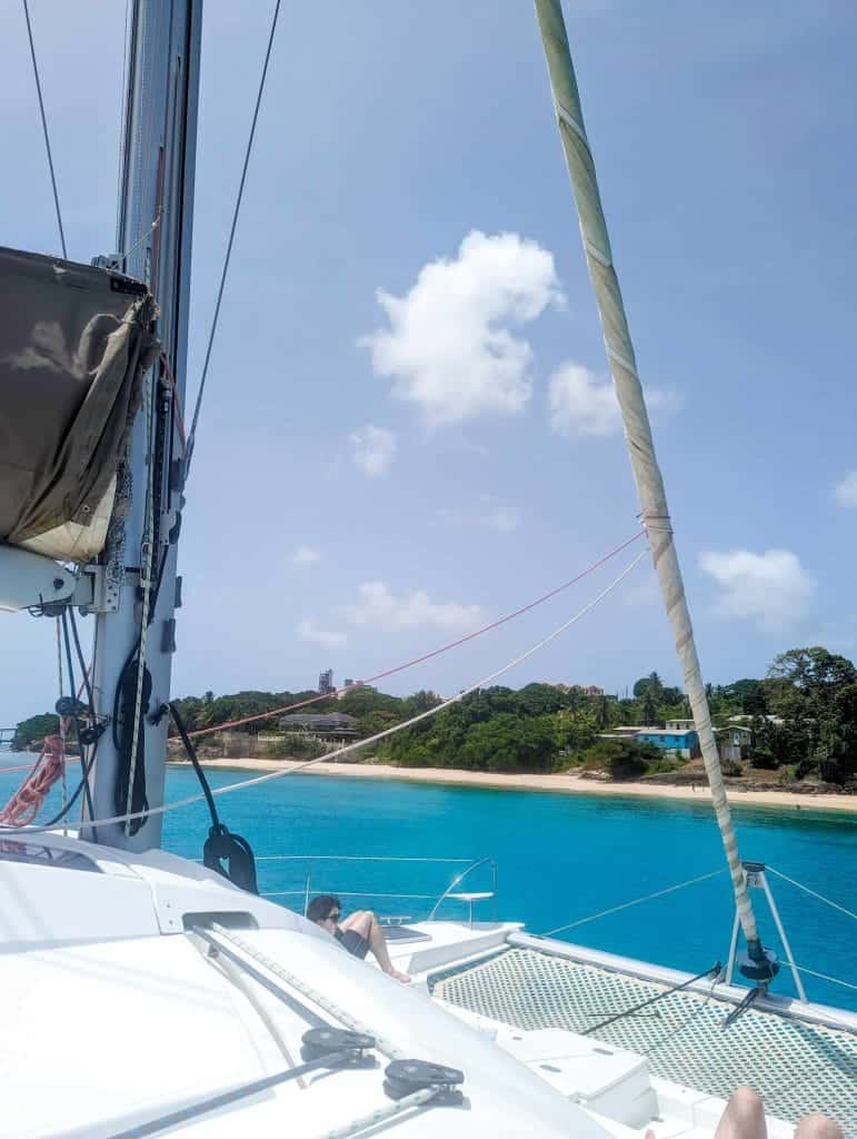 View from a sailboat deck with a sandy beach and lush greenery in the background under a clear blue sky.