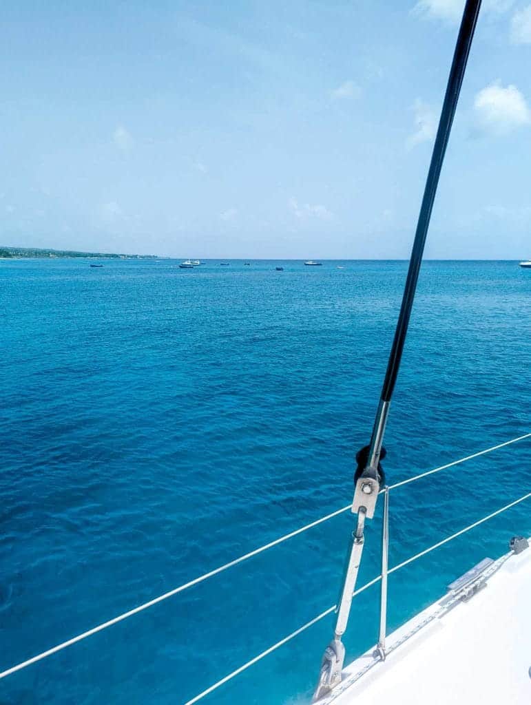 View from a sailboat showing a clear blue ocean and a few distant boats on the horizon.