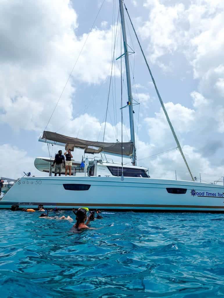 People snorkeling near a sailboat on clear blue water, with a partly cloudy sky in the background.
