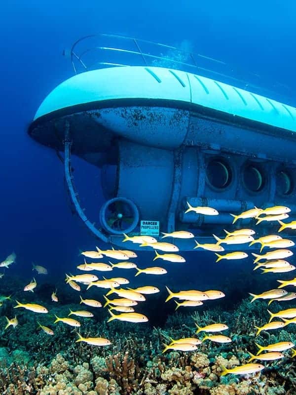 A submerged submarine near coral reef with a school of yellow fish swimming around it in clear blue water.