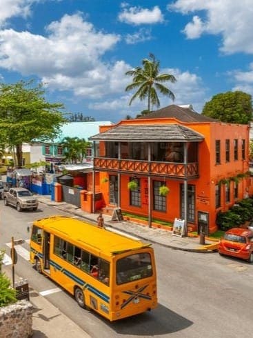 A vibrant orange building with a balcony stands on a sunny street with palm trees. A yellow bus drives by, with cars parked along the road and people walking on the sidewalk.