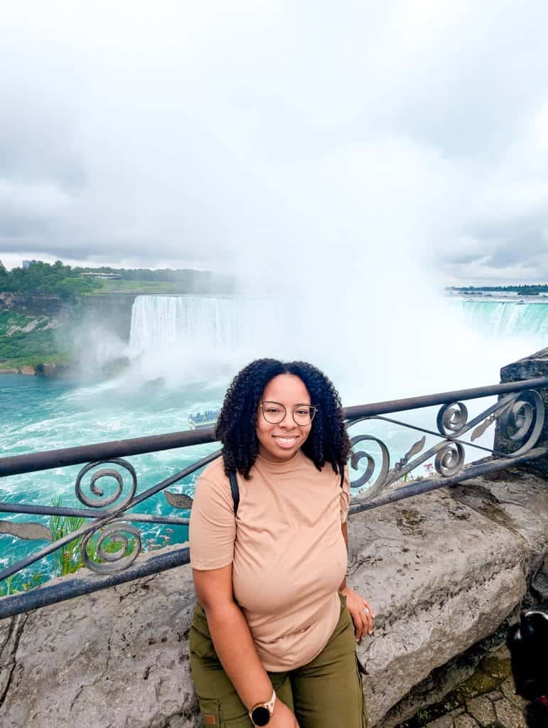 A person with curly hair and glasses is sitting on a stone ledge near a railing, checking off a bucket list experience as they admire the large waterfall in the background under a cloudy sky.