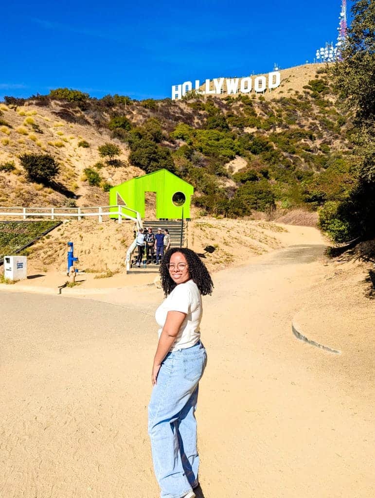 A person beams with joy in front of the iconic Hollywood Sign, a bucket list moment captured perfectly against the backdrop of clear blue skies and lush surrounding hills in Los Angeles.