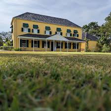 A yellow colonial-style house with green awnings sits on a grassy lawn under a cloudy sky.