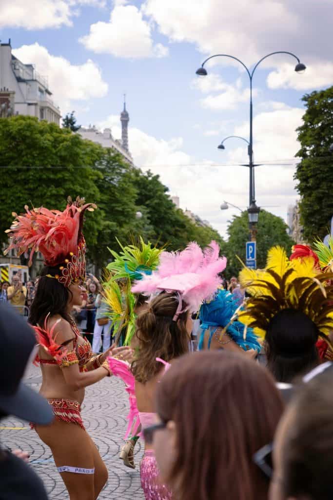 People in colorful feather costumes participating in a street parade with trees and a distant tower in the background.