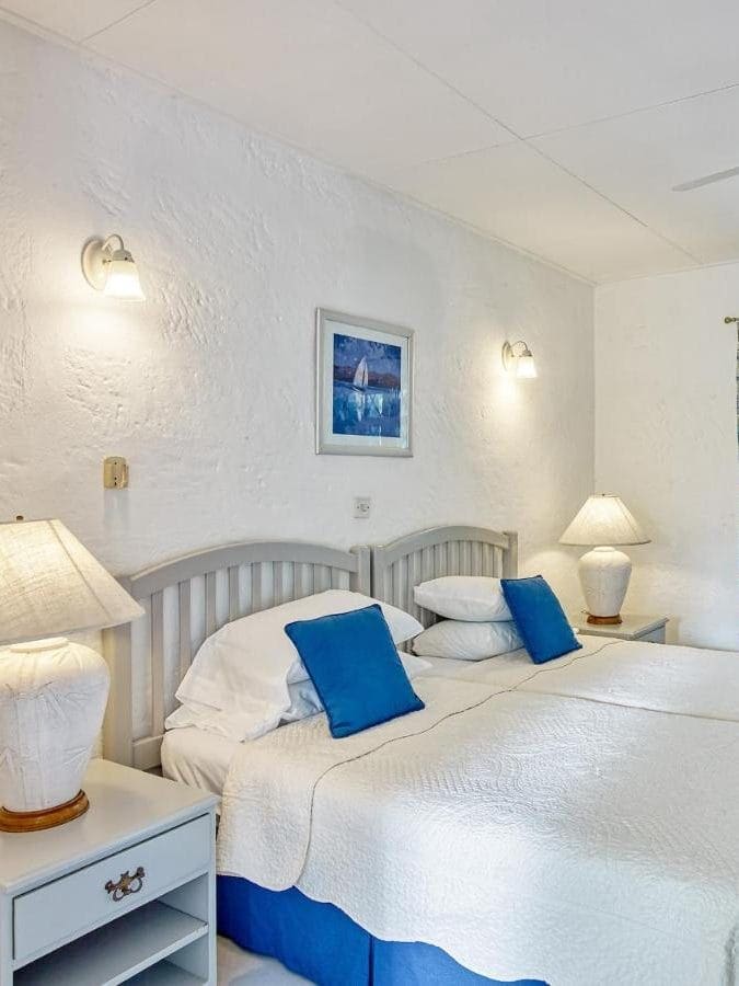 Bedroom with two single beds pushed together, white bedding, blue cushions, two bedside tables with lamps, a ceiling fan, and a window with patterned curtains.