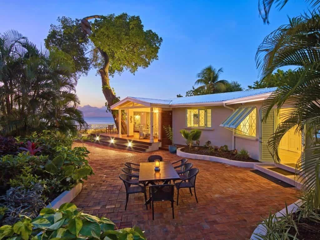 A cozy house with a patio and outdoor dining area surrounded by lush greenery and overlooking the ocean at sunset.