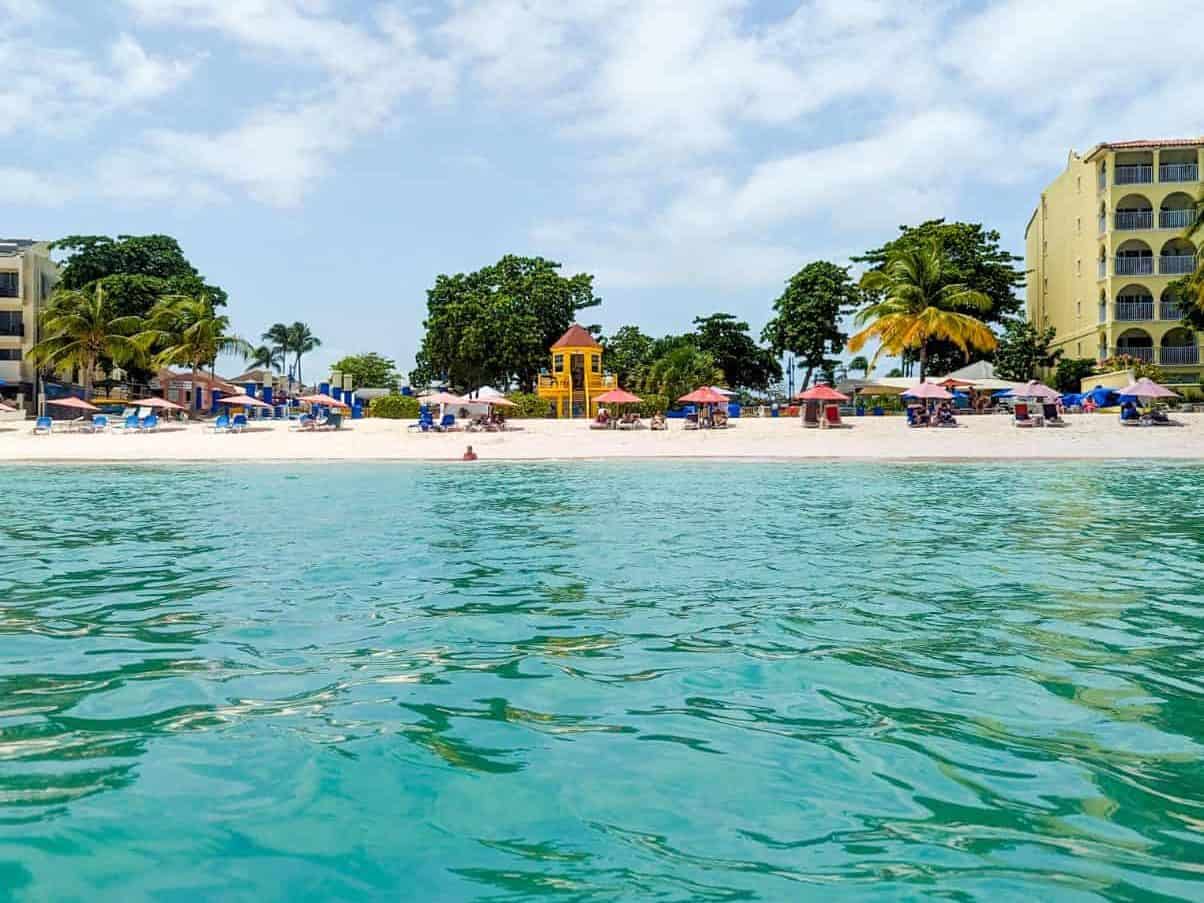 View of a sandy beach with sunbathers, umbrellas, palm trees, and buildings under a partly cloudy sky, seen from the turquoise ocean.