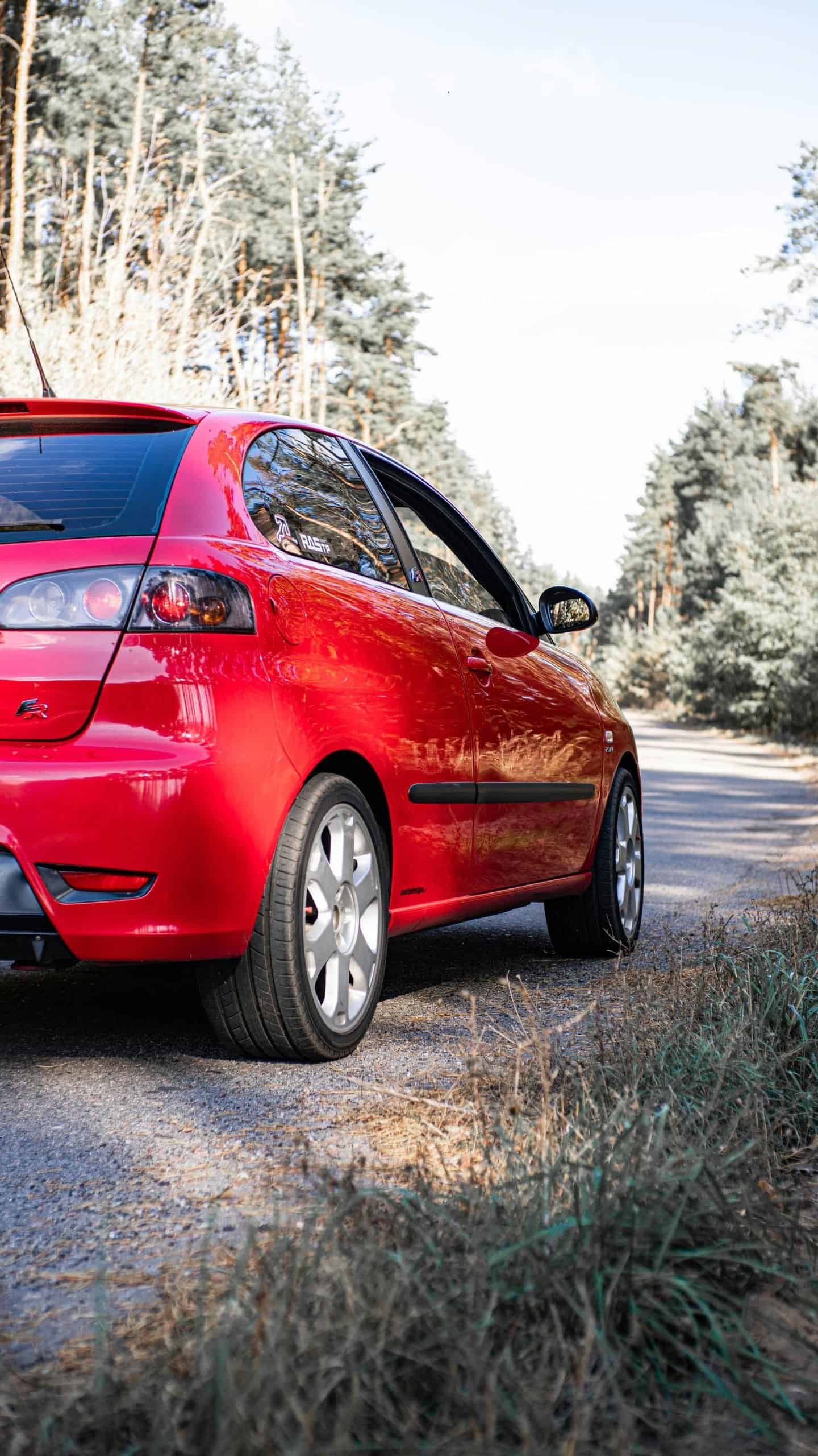 Red hatchback car parked on a rural road, surrounded by trees.