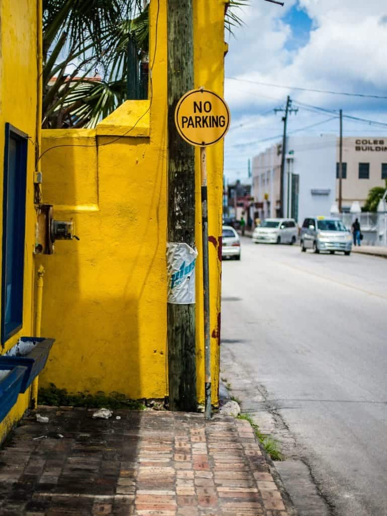 Street scene with a bright yellow wall and a "No Parking" sign on a utility pole. Cars are parked along the road under a blue sky.