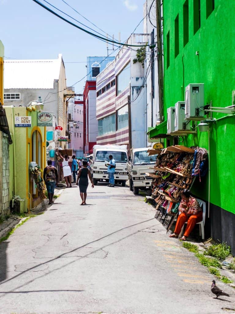 A narrow street with colorful buildings, parked vehicles, a shoe stall, and pedestrians walking.